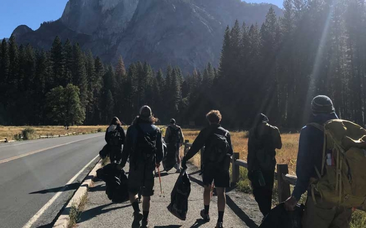 A group of outward bound students carry trash bags as they walk alongside a road during a service project. There are evergreen trees and a mountain in the background. 